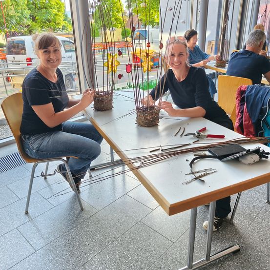 Zwei Frauen sitzen an einem Tisch mit Körben und Werkzeugen und lächeln. Sie scheinen mit Zweigen zu basteln. Hinter ihnen ist eine Glaswand, die einen Außenbereich mit Bäumen und einem Van zeigt.