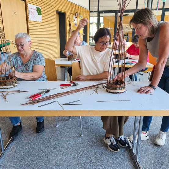 Mehrere ältere Frauen sind in einem Raum und arbeiten an Webprojekten an einem Tisch. Eine Frau leitet eine andere, und Werkzeuge sind auf dem Tisch verstreut.