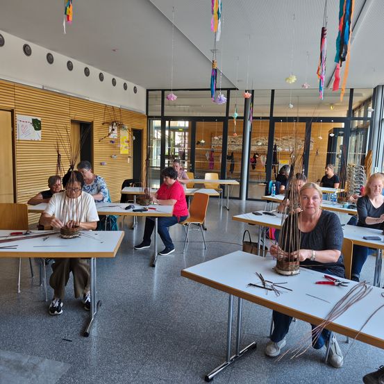 Eine Gruppe von Menschen sitzt in einem Workshop um Tische herum und bastelt mit Zweigen und Schnüren. Der Raum ist mit hängenden Dekorationen und Holzwänden gefüllt. Einige tragen eine Brille, andere haben Armbänder.