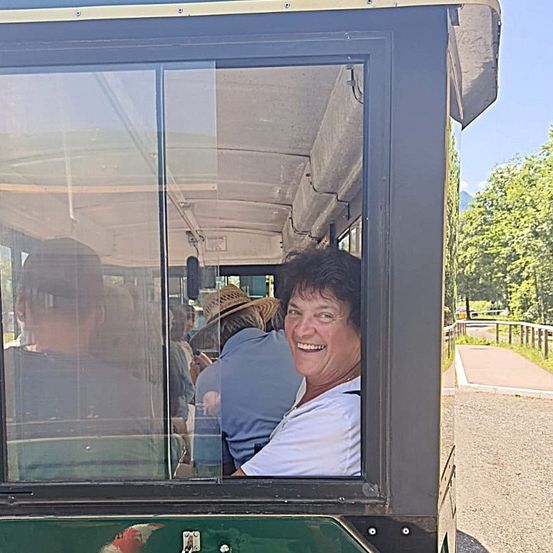 Eine Frau lächelt in einem Fahrzeug, sichtbar durch die Glasfenster. Andere Menschen sitzen hinter ihr, und das Fahrzeug ist auf einer Straße mit Bäumen und einem Zaun.