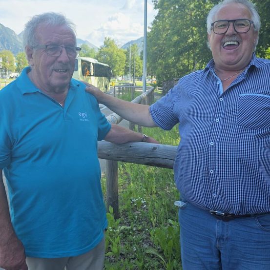 Zwei Männer stehen nebeneinander, einer trägt ein blaues Poloshirt, der andere ein kariertes Hemd, beide lächeln und posieren für ein Foto.