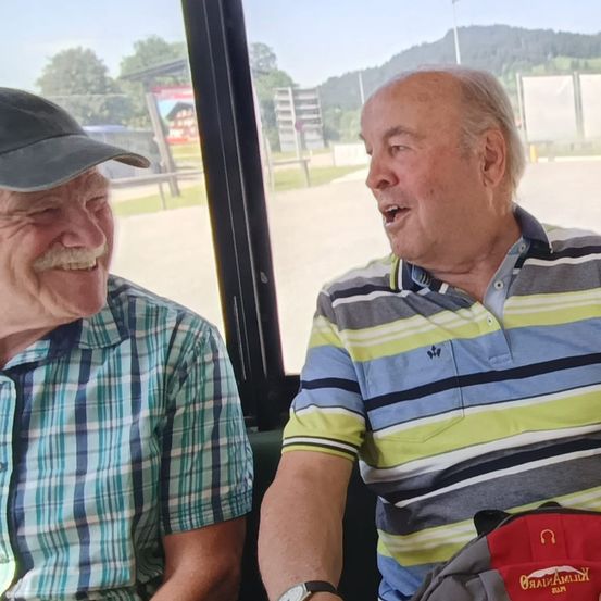 Zwei ältere Männer sitzen auf einem Bus, lächeln und schauen aus dem Fenster. Einer trägt ein kariertes Hemd, der andere ein gestreiftes Poloshirt. Sie sind von einer malerischen Aussicht umgeben.