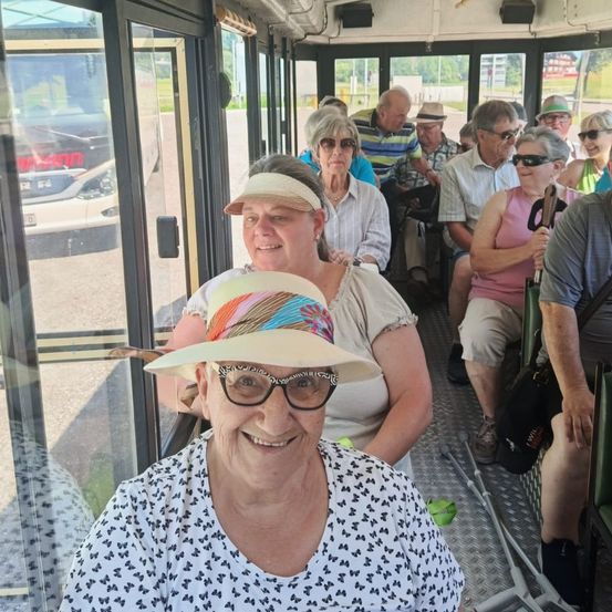Ein Bus mit mehreren älteren Leuten, die Hüte und Brillen tragen. Der Bus ist in einem Außenbereich geparkt. Die Leute im Inneren lächeln und scheinen eine Tour zu machen.