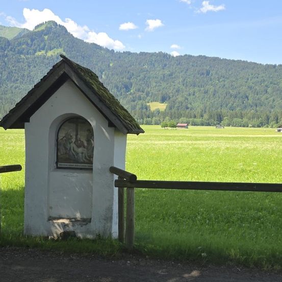 Eine kleine Kapelle mit einer religiösen Darstellung an der Vorderseite steht auf einer üppigen grünen Wiese, mit Bergen im Hintergrund.