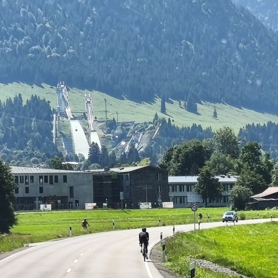 Eine Person fährt mit dem Fahrrad auf einer Bergstraße mit einem großen Gebäude im Hintergrund. Es gibt zwei Skisprungschanzen am Berg.