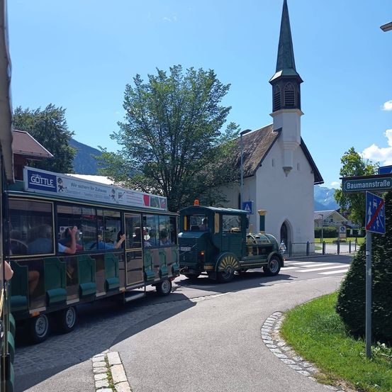 Zwei grüne Straßenbahnen fahren vor einer Kirche mit einem Kirchturm die Straße hinunter. Leute sitzen in den Straßenbahnen, einige machen Fotos. Ein Straßenschild befindet sich rechts.