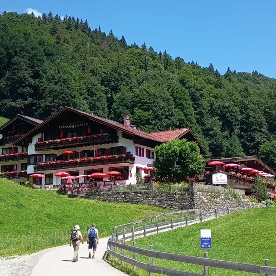 Zwei Wanderer mit Rucksäcken gehen einen Pfad entlang vor einem Gebäude mit roten Dächern und roten Sonnenschirmen, umgeben von saftigem grünem Gras und Bergen.