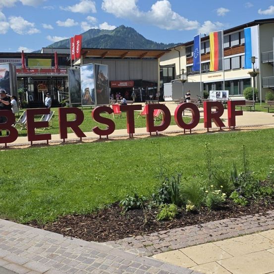 Ein rotes Schild mit dem Wort 'BERSTDORF' befindet sich auf einer Rasenfläche. Mehrere Menschen gehen in der Gegend herum. Im Hintergrund befinden sich Gebäude. Der Himmel ist blau mit einigen Wolken.