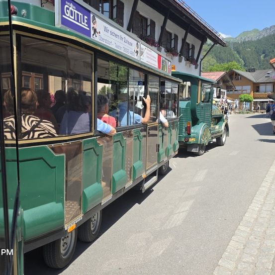 Touristen sitzen in einem grünen Trolley mit offenem Fenster auf einer asphaltierten Straße, mit Gebäuden und Bergen im Hintergrund.