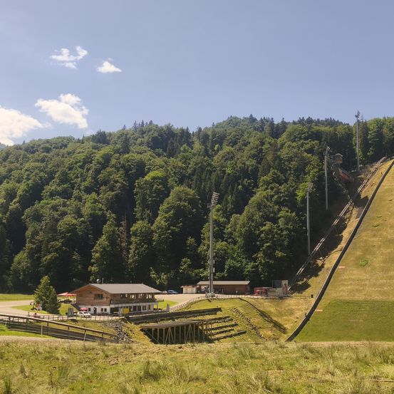 Ein Sprunghügel mit einer grün bewachsenen Anhöhe und einem Holzgebäude im Vordergrund. Der Hügel ist umgeben von üppigem grünen Bäumen unter einem blauen Himmel.