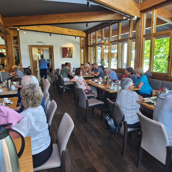 Eine Gruppe von Menschen sitzt an Tischen in einem mit Holz verkleideten Restaurant. Einige essen, während andere Gespräche führen. An der Wand hängt ein Bilderrahmen.