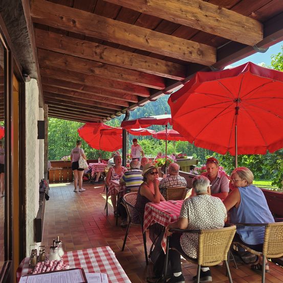 Eine Gruppe von Menschen sitzt mit roten Sonnenschirmen in einem Restaurant an Tischen. Die Tische sind mit rot-weiß karierten Tischdecken bedeckt. Das Restaurant hat Holzwände und -böden.