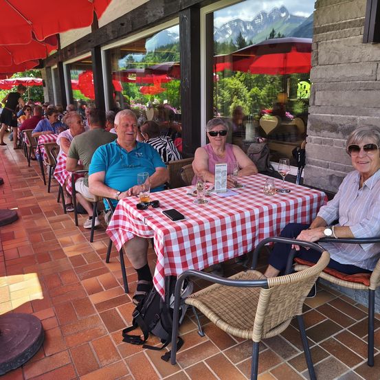 Ein Outdoor-Café mit Gästen, die an Tischen unter roten Schirmen sitzen. Berge sind im Hintergrund durch die Glasfenster sichtbar.
