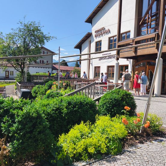 Kurhaus Fiakina Restaurant mit Sitzgelegenheiten im Freien. Leute stehen vor dem Eingang, einige gehen. Das Gebäude hat Glasfenster und einen Balkon.
