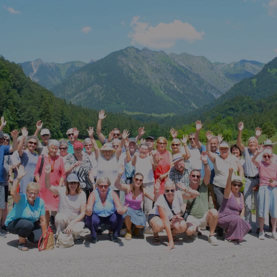 Eine Gruppe von Menschen in einem Gebirgsgebiet lächelt und posiert für ein Foto. Sie tragen alle Sonnenbrillen und einige winken. Die Berge sind mit Bäumen und Pflanzen bedeckt.