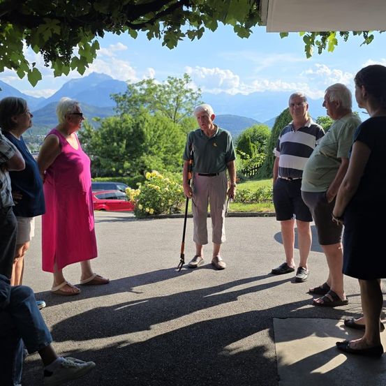 Eine Gruppe von Menschen steht auf einem Parkplatz und unterhält sich, mit Bergen und Bäumen im Hintergrund. Eine Frau in Pink steht in der Nähe eines Mannes mit Gehstock.