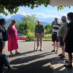 Eine Gruppe von Menschen steht auf einem Parkplatz und unterhält sich, mit Bergen und Bäumen im Hintergrund. Eine Frau in Pink steht in der Nähe eines Mannes mit Gehstock.