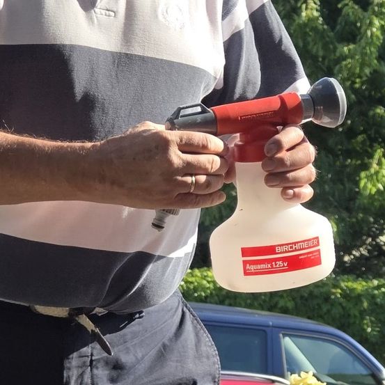 Eine Person hält eine rot-weiße Sprühflasche mit dem Text Birchmeier Aquamix 1,25V. Die Person steht vor einem Auto.
