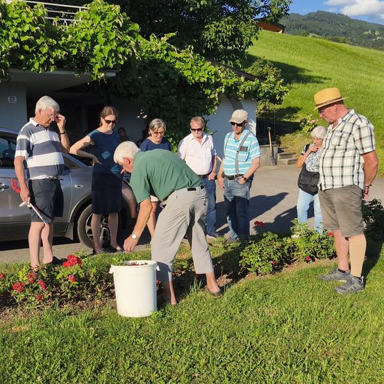 Eine Gruppe von Menschen versammelt sich in einem Garten, einer Mann pflückt Blumen. Die Gruppe steht in der Nähe eines Autos, mit einem Gebäude und Bergen im Hintergrund.