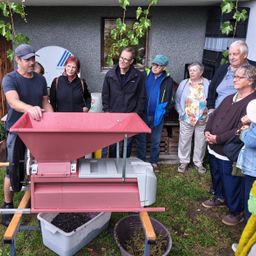A group of people are gathered around a red machine, possibly a grape crusher, in a backyard. They are observing the process while a man in a blue shirt is operating it.
