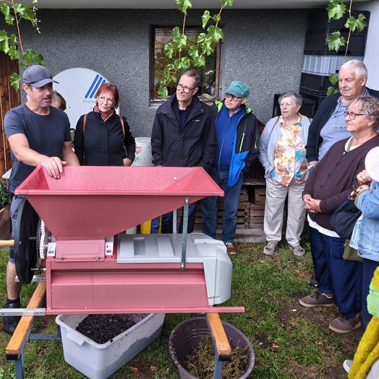 Eine Gruppe von Menschen steht um eine rote Maschine herum, wahrscheinlich einen Traubenpresser, im Garten. Sie beobachten den Prozess, während ein Mann in einem blauen Shirt sie bedient.