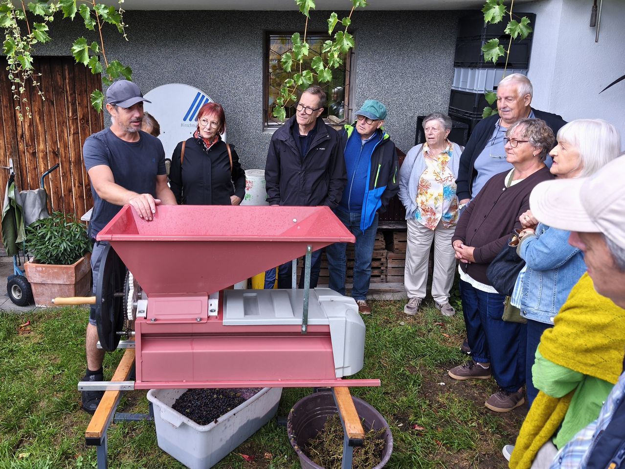 Eine Gruppe von Menschen steht um eine rote Maschine herum, möglicherweise eine Traubenpresse, in einem Hinterhof. Die Maschine ist mit Trauben gefüllt.