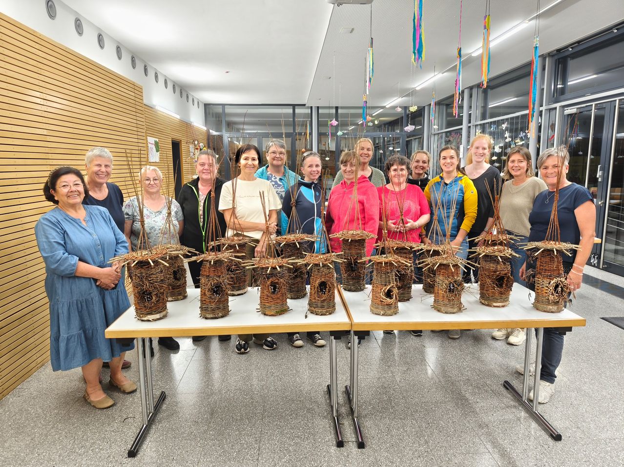 Eine Gruppe von Menschen steht lächelnd in einem Raum. Sie halten kleine, gewebte Körbe. Vor ihnen stehen Tische mit weiteren Körben. Der Raum hat Holzwände und Glasfenster.