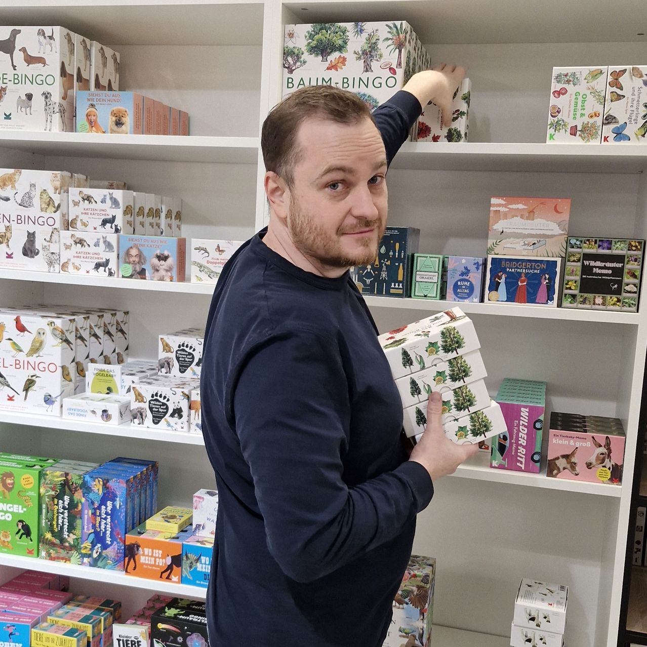 Ein Mann, der eine Schachtel mit Karten hält, steht vor einem weißen Regal voller verschiedener Spiele und Schachteln.
