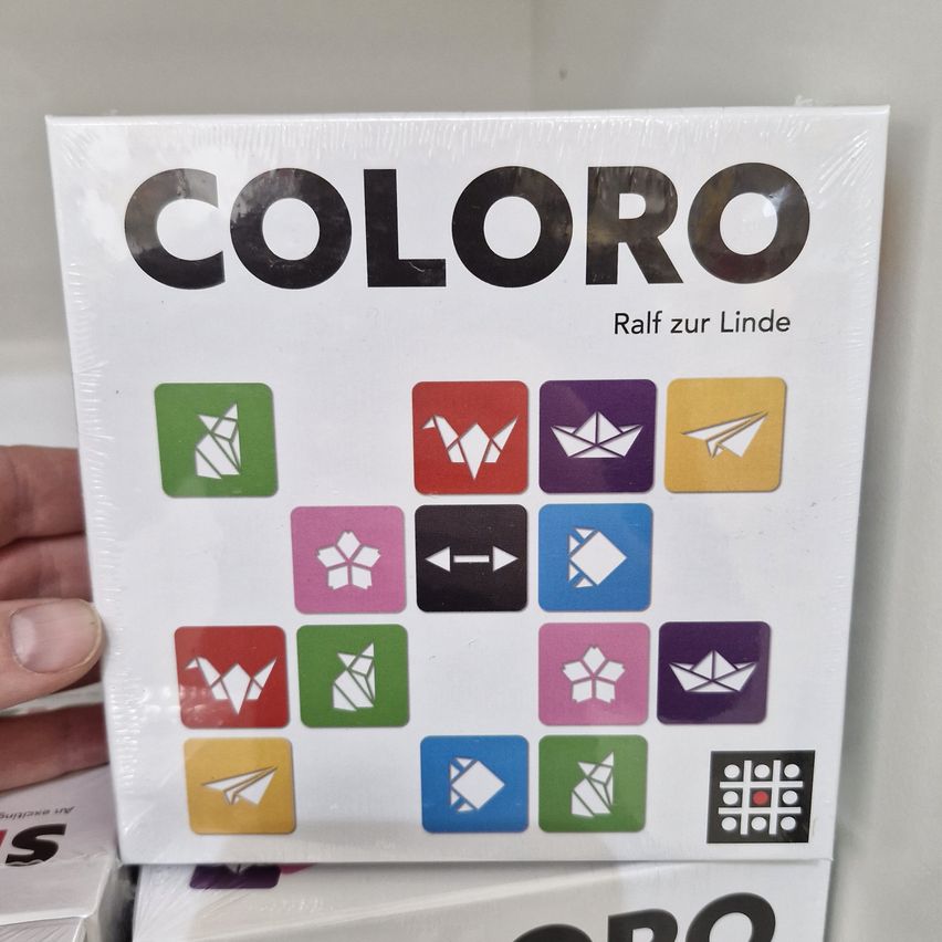 Eine Schachtel des Brettspiels 'Coloro' wird gezeigt, mit verschiedenen farbigen Symbolen und der Phrase 'Raff zur Linde' darauf. Die Schachtel wird von einer Hand gehalten.