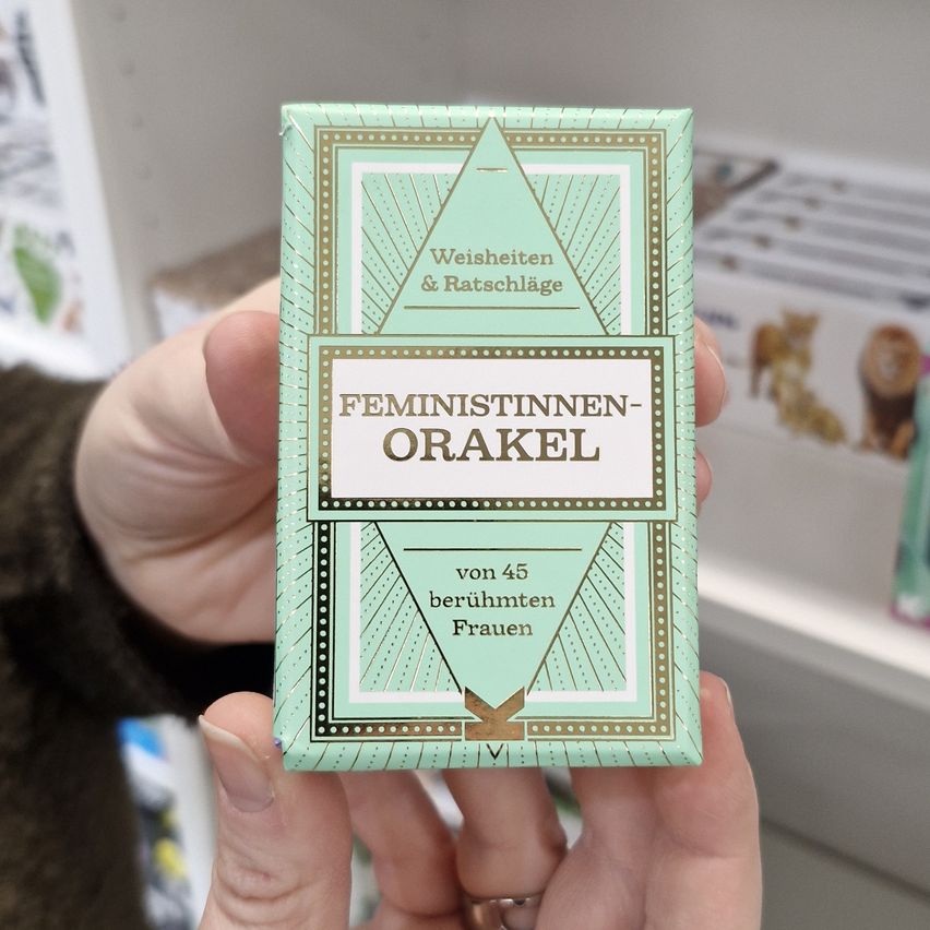 Eine Person hält eine kleine Schachtel mit der Aufschrift 'Feministinnen-Orakel', die einen grünen Deckel mit geometrischem Design hat. Die Schachtel ist mit Karten für das Wahrsagen gefüllt.
