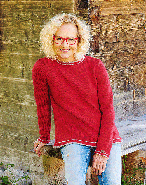 Eine Frau mit blonden lockigen Haaren, die einen roten Pullover und blaue Jeans trägt, sitzt auf einer Holzbank vor einer Holzwand.
