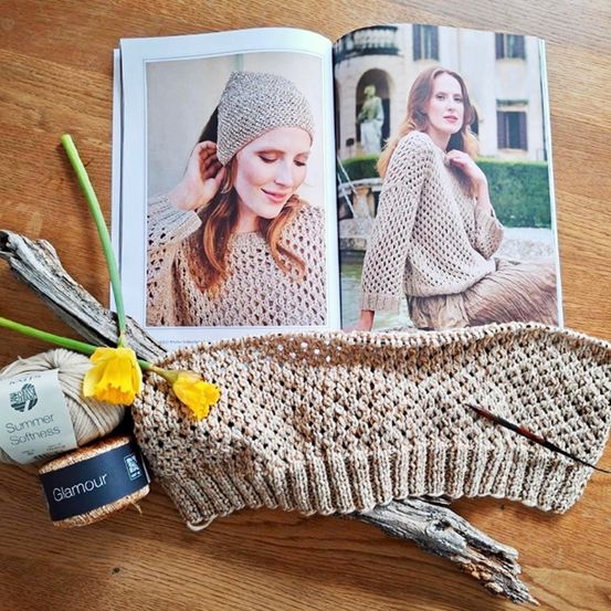 Ein offenes Magazin mit Häkelanleitungen, einem gestrickten Mützchen und Garn auf einem Holztisch.
