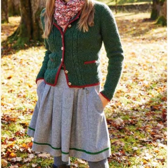 Eine Frau trägt einen grünen Cardigan mit rotem Besatz und einem gemusterten Schal. Sie steht in einem Grasbereich mit verstreuten Blättern.