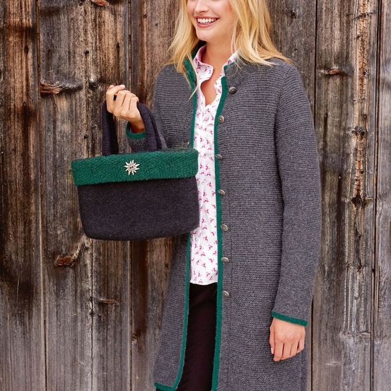 Eine Frau steht vor einer Holzwand und hält einen grauen und grünen gestrickten Cardigan mit einem dekorativen Stern und einer passenden Handtasche.