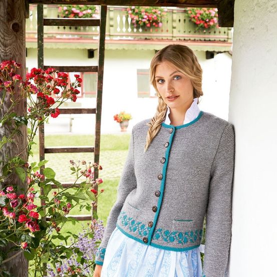 Eine Frau in einem grauen Strickjacke mit türkisfarbenem Rand und einem blauen Kleid steht an einer Wand mit Blumen. Im Hintergrund befindet sich ein Haus mit einem Balkon, das mit rosa Blumen geschmückt ist.