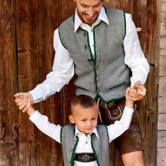 Ein Mann in einer grauen Weste mit grünem Besatz und einem weißen Hemd hält die Hände eines Jungen, der ein traditionelles Outfit trägt.