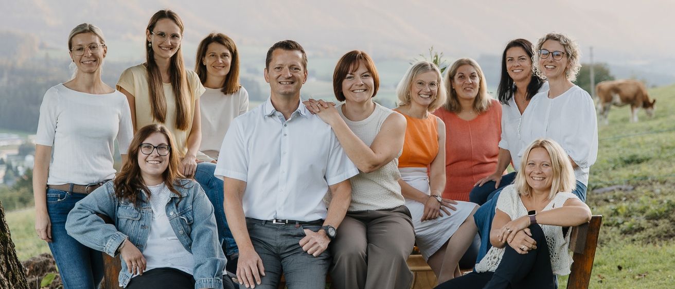 Eine Gruppe von Menschen posiert für ein Foto auf einer Bank unter einem Baum. Sie lächeln und blicken in verschiedene Richtungen. Hinter ihnen grast eine Kuh auf einem grasigen Feld. Der Hintergrund zeigt einen Hügel unter einem klaren Himmel.