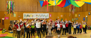 Eine Gruppe von Kindern führt ein Theaterstück in einer Turnhalle auf, trägt verschiedene Kostüme und Masken. Bunte Dekorationen hängen von der Decke, und das Publikum sitzt im Vordergrund.