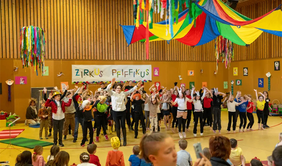 Eine Gruppe von Kindern führt ein Theaterstück in einer Turnhalle auf, trägt verschiedene Kostüme und Masken. Bunte Dekorationen hängen von der Decke, und das Publikum sitzt im Vordergrund.