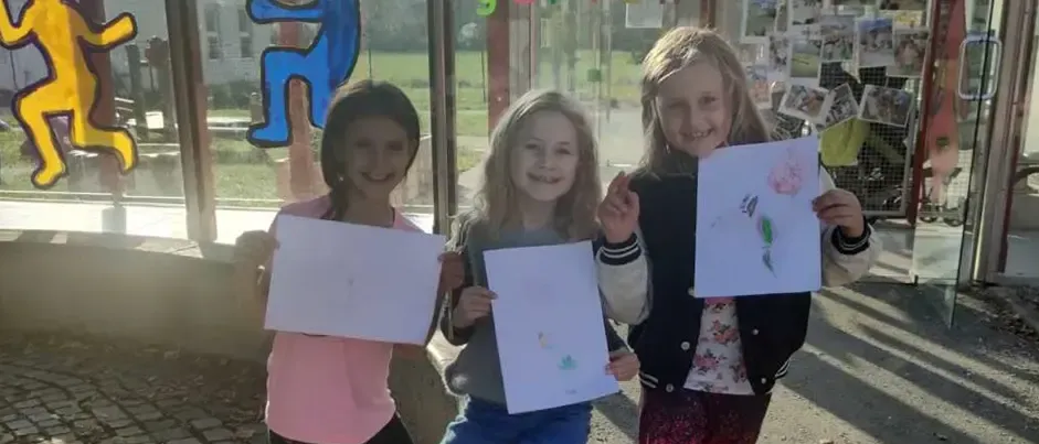 Drei junge Mädchen lächeln und halten ihre Zeichnungen vor einem Kindergartengebäude hoch. Sie stehen neben einer Bank mit Malutensilien.
