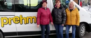 Drei Frauen stehen vor einem weißen Van mit der Aufschrift 'prehm' und lächeln für ein Foto. Dahinter befinden sich Häuser mit gelben Wänden und eine Straße.