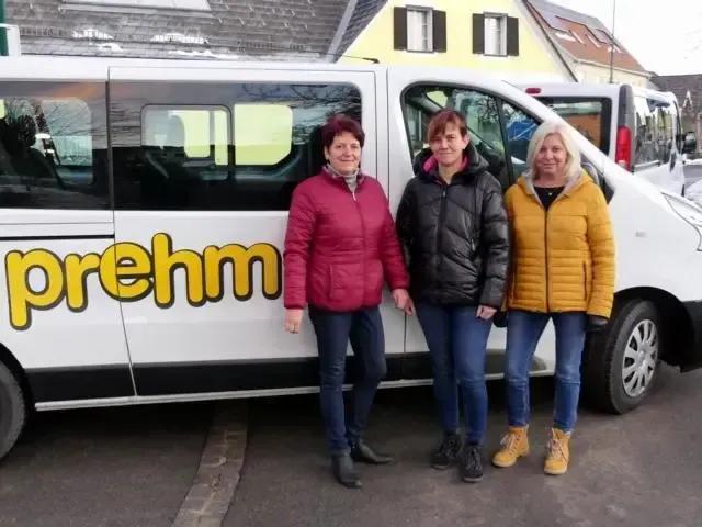 Drei Frauen stehen vor einem weißen Van mit der Aufschrift 'prehm' und lächeln für ein Foto. Dahinter befinden sich Häuser mit gelben Wänden und eine Straße.