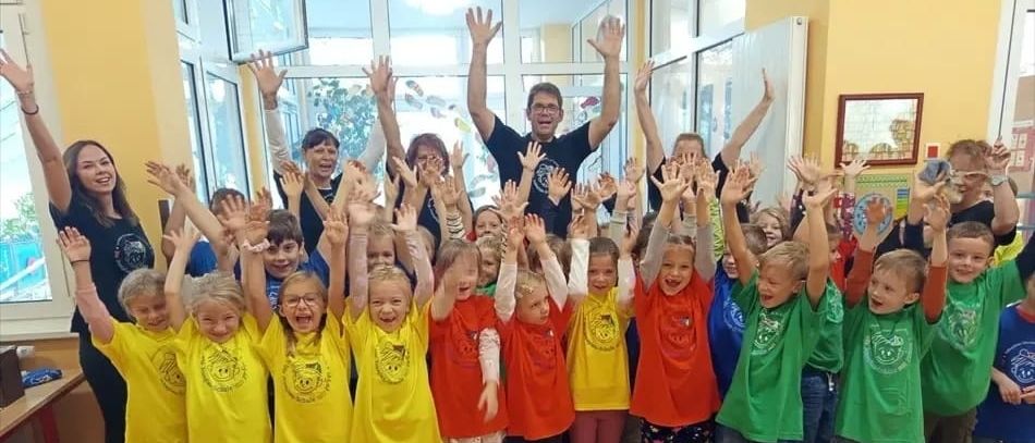 Gruppenfoto von Kindern und Erwachsenen, die bunte T-Shirts tragen, in einem Raum mit erhobenen Händen stehen, in der Nähe von Fenstern und einer Tür.