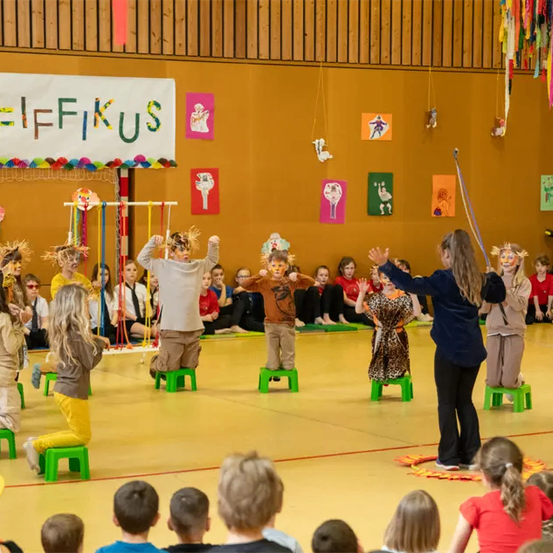 Eine Gruppe von Kindern in Kostümen führt mit einer Lehrerin eine Aufführung auf der Bühne auf, während Zuschauer zuschauen. Die Bühne hat ein Banner mit der Aufschrift 'Pfifikus'.