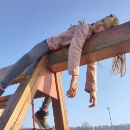 Ein kleines Mädchen in einem rosa Langarmshirt und Jeans hängt an einer Holzstruktur, möglicherweise einem Spielplatz. Sie schaut mit einem Lächeln nach oben.