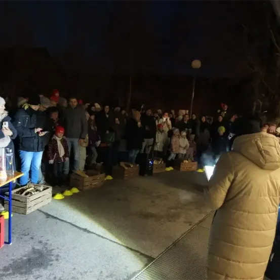 Eine Menschenmenge in Winterkleidung versammelt sich bei Nacht im Freien. Sie stehen um Holzkisten und einen Tisch mit Flaschen. Eine Person mit Hut und Mantel steht im Vordergrund.