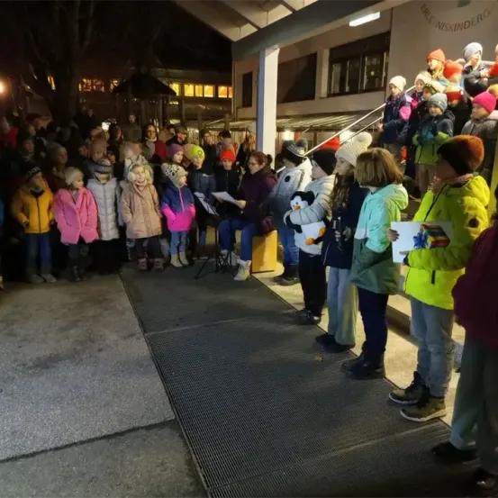 Eine Gruppe von Kindern und Erwachsenen steht vor einem Gebäude mit einem Schild, auf dem Eke Niske steht. Sie sind warm gekleidet in Mänteln und Mützen. Einige halten Papiere in der Hand und ein Kind hält einen weichen Pinguin.