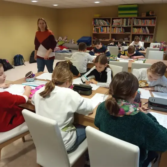 In einem Klassenzimmer sitzen mehrere Kinder an Tischen und arbeiten an ihren Aufgaben. Eine Lehrerin steht in der Nähe eines Bücherregals und leitet sie. Der Raum ist gut beleuchtet, mit verschiedenen Utensilien und Büchern in der Umgebung.