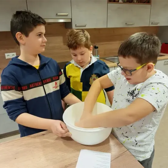 Drei Jungen sind in einer Küche, einer rührt etwas in einer Schüssel, während die anderen zusehen. Sie befinden sich in einem Raum mit Holzschränken und einer Spüle.