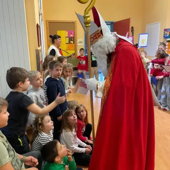 Ein Mann, der als Bischof verkleidet ist, interagiert mit Kindern in einem Klassenzimmer. Die Kinder sitzen auf dem Boden, und einige stehen. Der Raum hat gelbe Wände und Holzböden.